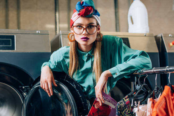 woman in glasses and turban holding magazine near modern washing machine in public laundromat woman in glasses and turban holding magazine near modern washing machine in public laundromat turban stock pictures, royalty-free photos & images