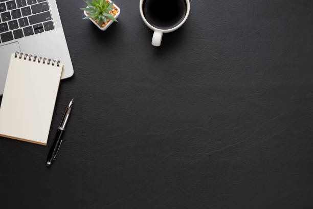 istock Top view above of Black leather office desk table with keyboard, notebook and coffee cup with equipment office supplies. Business and finance concept. Workplace, Flat lay with blank copy space. 1284056129