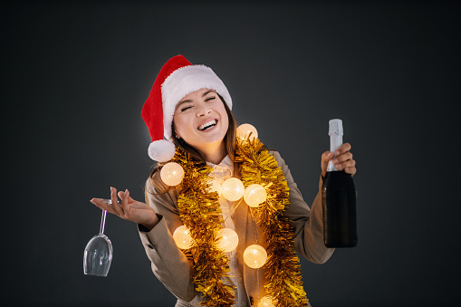 Young smiling businesswoman with santa's cap and christmas decorations around neck holding champagne and glass on christmas eve in front of gray background.