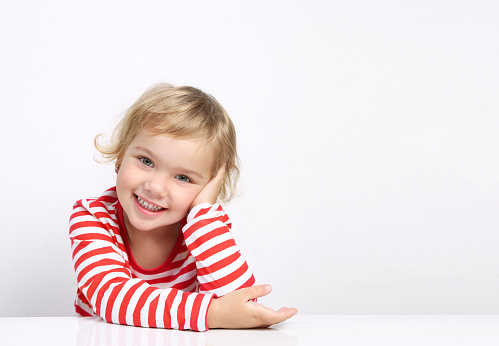 Caucasian child portrait smiling face sitting at empty space table on white background.Advertisement design,promotion.Kid girl shows copy space.