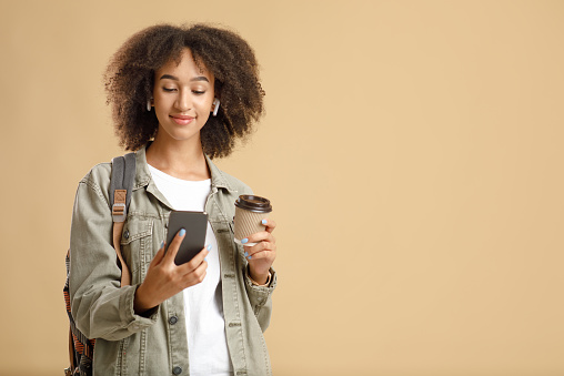 Watch movie, read blog, get message and active lifestyle. Friendly calm african american woman looking on smartphone holds cup of takeaway coffee isolated on light background, copy space, studio shot
