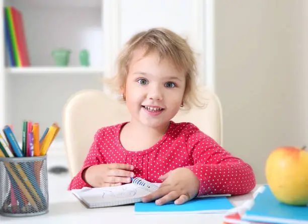 Little smiling girlsitting at table and drawing in notebook.Prescooler education,home learning.Caucasian kid studies.