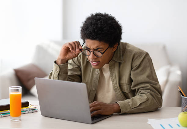 Poor eyesight. Black teenager in glasses squinting his eyes while using laptop computer for online education at home Poor eyesight concept. Black teenager in glasses squinting his eyes while using laptop computer for online education at home. African American youth having vision problem because of PC overuse squint eyes stock pictures, royalty-free photos & images