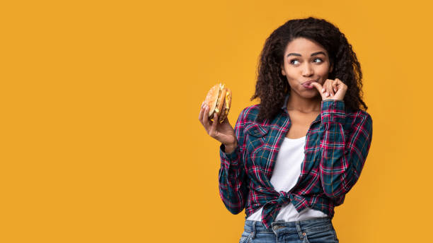Happy African American Lady Holding Burger At Studio Cheat Meal. Portrait Of Happy African American Casual Woman Eating Tasty Burger, Licking Finger, Looking Away At Copy Space. Satisfied Woman Holding Fastfood, Isolated Over Orange Background, Banner licking stock pictures, royalty-free photos & images