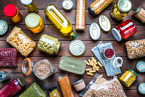 Top view of a large group of multicolored non-perishable canned goods, conserves, sauces, cereals, beans, nuts, crackers and oils scattered a wooden table. The composition includes cooking oil bottle, pasta, beans, preserves and tins. High resolution 42Mp studio digital capture taken with SONY A7rII and Zeiss Batis 40mm F2.0 CF lens