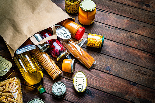 High angle view of a large group of multicolored non-perishable canned goods, conserves, sauces, crackers, cereals, beans, and oils coming out a brown paper bag shot on wooden table. The composition includes cooking oil bottle, pasta, beans, preserves and tins. High resolution 42Mp studio digital capture taken with SONY A7rII and Zeiss Batis 40mm F2.0 CF lens