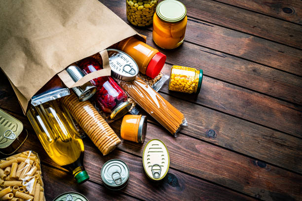 Non-perishable foods coming out a paper bag. Copy space High angle view of a large group of multicolored non-perishable canned goods, conserves, sauces, crackers, cereals, beans, and oils coming out a brown paper bag shot on wooden table. The composition includes cooking oil bottle, pasta, beans, preserves and tins. High resolution 42Mp studio digital capture taken with SONY A7rII and Zeiss Batis 40mm F2.0 CF lens canned food stock pictures, royalty-free photos & images