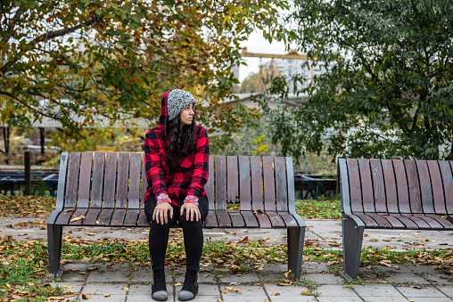 Young depressed homeless girl or woman with hat sitting alone on the bench on the street in the cold weather feeling desperate and anxious selective focus