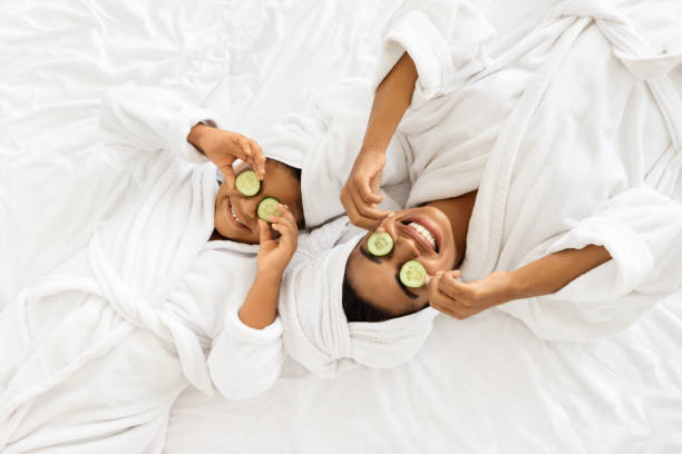 Black Mom And Daughter In Bathrobes Lying With Cucumber Slices On Eyes Beauty Spa Day. Cheerful Black Mom And Daughter In Bathrobes Lying With Cucumber Slices On Eyes, Doing Face Mask Treatment, Wearing Towel On Head, Having Fun Together At Home, Top View With Copy Space girls bath stock pictures, royalty-free photos & images