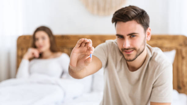 concentre-se na pílula azul nas mãos de um jovem feliz, e esposa sorridente senta-se na cama no interior moderno do quarto - bedroom authority bed contemporary - fotografias e filmes do acervo