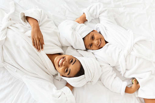 Top View Of Happy Black Mother And Daughter In Bathrobes And Towels Lying On Bed Relaxing After Spa, Pampering Themself With Beauty Treatments At Home, Mom And Child Having Fun Together, Free Space