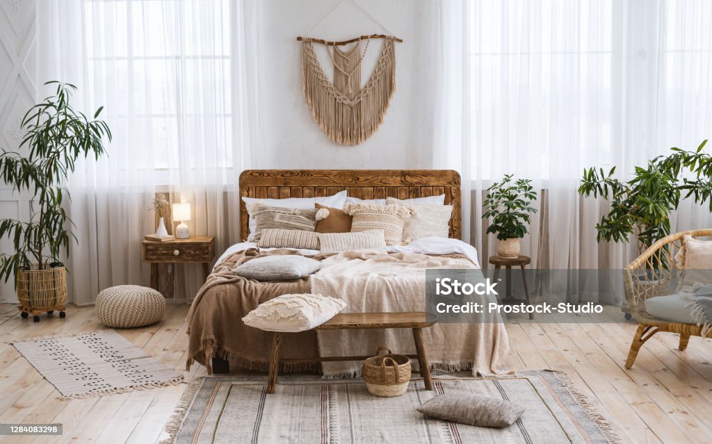 Rustic home design with ethnic boho decoration. Bed with pillows, wooden furniture Rustic home design with ethnic decoration. Bed with pillows, wooden furniture, plants in pots, armchair and curtains on large windows in cozy bedroom interior, nobody, flat lay, panorama, free space Bedroom Stock Photo