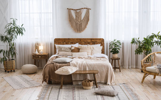 Rustic home design with ethnic boho decoration. Bed with pillows, wooden furniture Rustic home design with ethnic decoration. Bed with pillows, wooden furniture, plants in pots, armchair and curtains on large windows in cozy bedroom interior, nobody, flat lay, panorama, free space rustic stock pictures, royalty-free photos & images