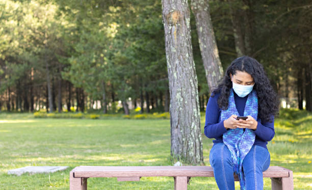 istock young woman dressed in blue and wearing mask, sitting on a bench in a park, while checking her cell phone 1284089747