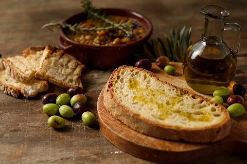 Composition with extra virgin olive oil on homemade bread on cutting board with legume soup and croutons of bread as a background together with glass ampoule with oil.