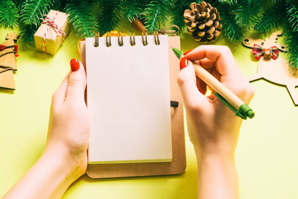 istock Top view of female hand making some notes in noteebok on yellow background. New Year decorations and toys. Christmas time concept 1284098262