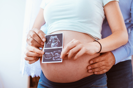 Pregnant couple of husband and wife feels love and relax at home. Young expecting woman holds baby in pregnant belly. Father take care of pregnant mother. Concept of maternity and pregnancy care. They holding ultrasound image
