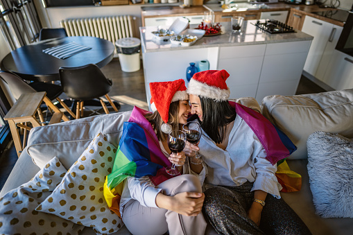 Photo of two young happy girlfriends sitting on the bed, wrapped in a lgbt flag and toasting on New Years Eve.