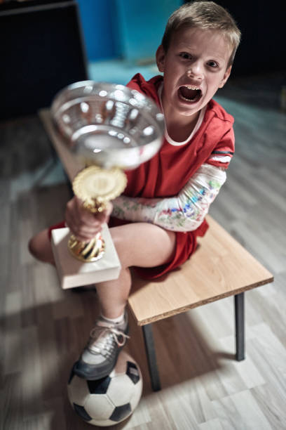 pequeño jugador emocionado posando con el trofeo ganado en el partido. deporte de equipo infantil - physical injury sport child locker room fotografías e imágenes de stock