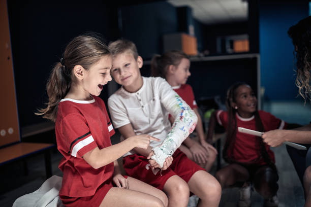 un pequeño jugador con un brazo roto en yeso mostrando su lesión a un compañero de equipo. deporte de equipo infantil - physical injury sport child locker room fotografías e imágenes de stock