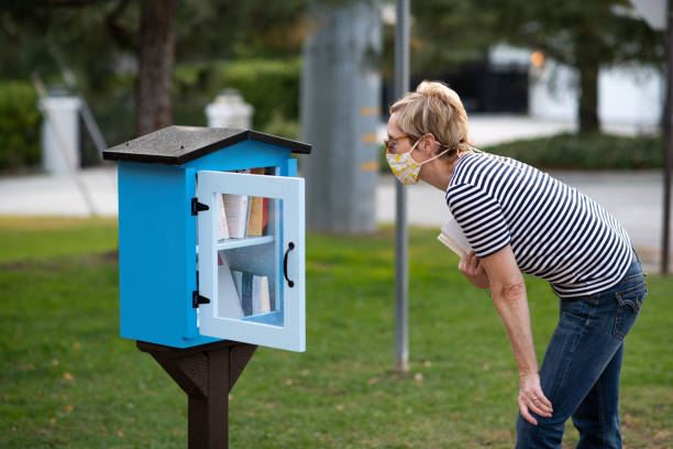 Female tourist mid fifties with face mask selecting a book from a free share a book mini library in Glenola Park at 5:30PM Pacific time. This charming park is located in LA Canada, California, USA. The sleepy town of LA Canada is a suburb of Los Angeles.
