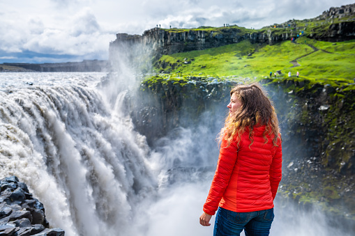 Young woman back looking at Dettifoss waterfall on rocks water flowing mist spraying cloudy day in Iceland with orange jacket and jeans