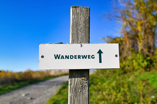 Sign on wooden pole saying 'hiking trail' in German with blurry landscape with blue sky in background