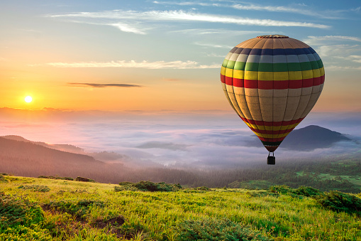 Big hot air baloon over idyllic landscape with green grass covered morning mountains with distant peaks and wide valley full of thick white cloudy fog.