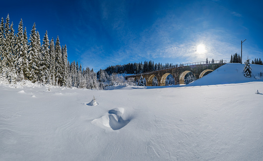 Stone viaduct (arch bridge) on railway through mountain snowy fir forest. Snow drifts  on wayside and hoarfrost on trees and electric line wires.