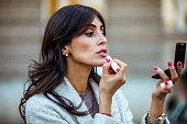 Beautiful mid adult woman holding red lipstic and doing quick makeup on the city street