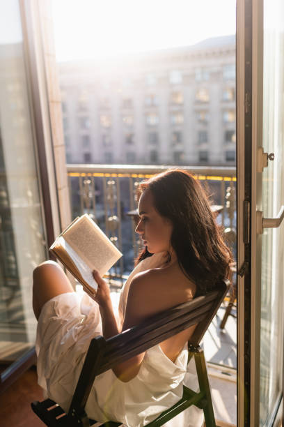 attractive woman covered in white sheet reading book and sitting on balcony