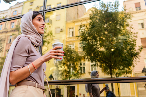 Smiling Muslim woman wearing hijab headscarf walking in the city center. Arab female in urban background