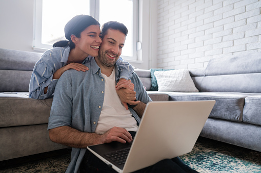 Young Caucasian man sitting on the floor and using a laptop while his girlfriend is hugging him. Happy young Caucasian couple using a laptop together at home.