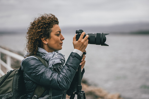 Young female photographer with backpack standing on riverbank and taking a photo of river with digital camera, side view