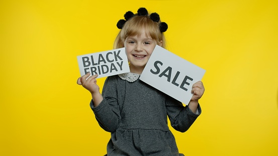 Attractive joyful young child girl showing Black Friday and Sale word inscriptions notes, smiling, looking satisfied with low prices, shopping on holiday. Preschooler kid rejoicing good discounts