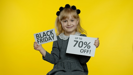 Little joyful blonde child girl showing Black Friday and Up To 70 Percent Off inscriptions, smiling, looking satisfied with low prices, shopping on holiday. Preschooler kid rejoicing good discounts