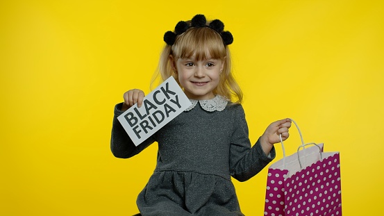Cute happy little child girl with shopping bag showing Black Friday inscription, smiling, looking satisfied with low prices, discounts purchases. Pupil teen kid on yellow background
