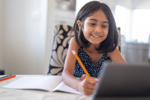 chica linda de la edad primaria usando computadora portátil mientras asiste a la escuela en línea - educación en el hogar fotografías e imágenes de stock