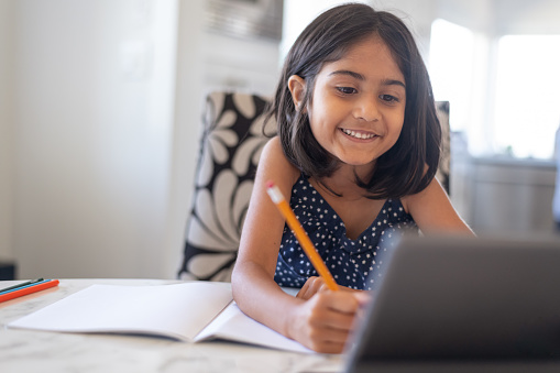 An adorable elementary age girl of Indian descent smiles while doing school assignment online as she studies at home during Covid-19 pandemic. The girl is using a tablet computer to attend a class using a video call. Distance learning and homeschooling concepts.