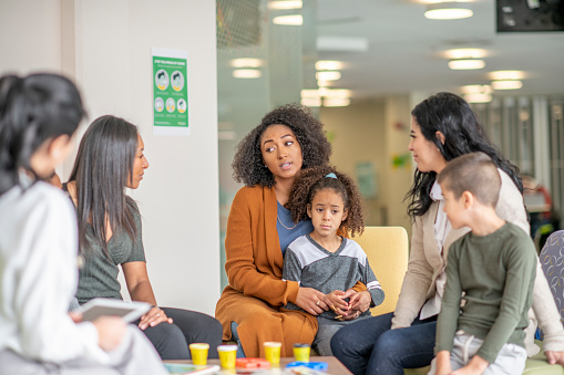 Women and kids sit around a table in a colourful facility and discuss.