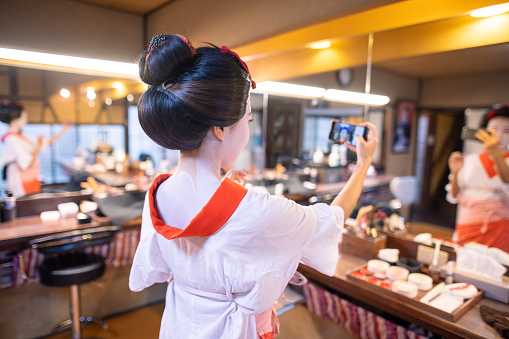 Young female tourist visiting Kyoto in Japan and experiencing Maiko (Geisha in training) makeover. Applying special white face makeup before wearing traditional Japanese Maiko kimono.