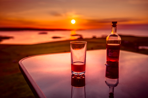A bottle of gin liqueur and one glass on the table with a beautiful sunset in the background, Unst, the northernmost island in Great Britain.