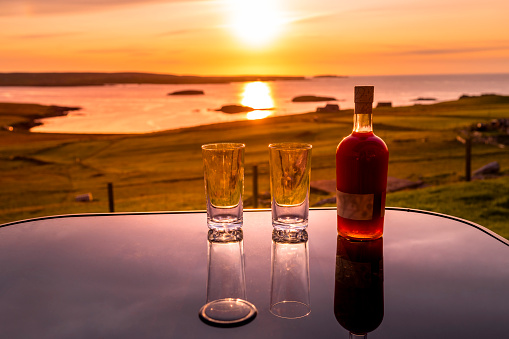 A bottle of gin liqueur and two glasses on the table with a beautiful sunset in the background, Unst, the northernmost island in Great Britain.