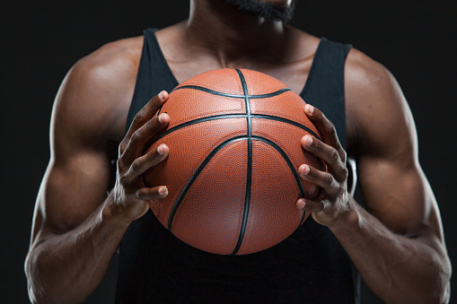 Portrait of afro american male basketball player playing with a ball over black background. Fit young man in sportswear holding basketball.