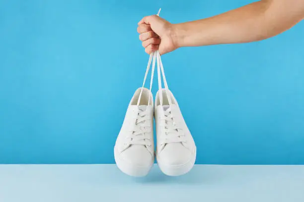 The hand holds by shoelaces Pair of fashion stylish white sneakers on a pastel blue background.