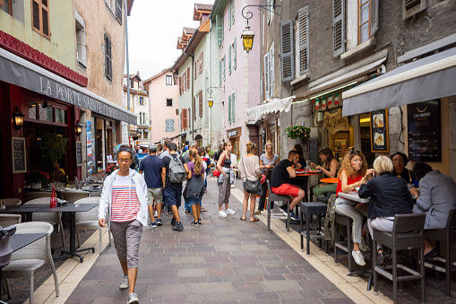 This is a street near the river in Annecy, France. It is overcast after a rain shower there people eating at local restaurants and an open area of colourful buildings and tourists walking past.
