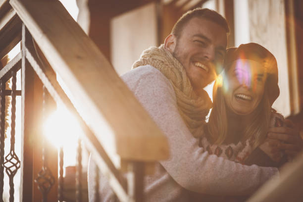 Cheerful young man embracing and cuddling his girlfriend while they are having a laugh and sitting outdoors in warm winter sunshine on their weekend getaway at the rustic cabin.