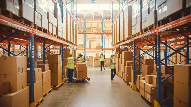 Photo of Retail Warehouse full of Shelves with Goods in Cardboard Boxes, Workers Scan and Sort Packages, Move Inventory with Pallet Trucks and Forklifts. Product Distribution Delivery Center.