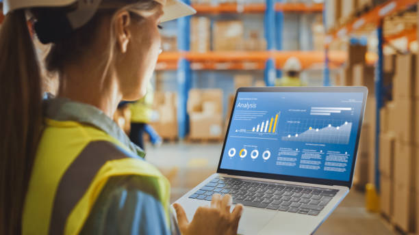 istock Professional Female Worker Wearing Hard Hat Holds Laptop Computer with Screen Showing Analysis Software in the Retail Warehouse full of Shelves with Goods. Over the Shoulder Side View 1284193231