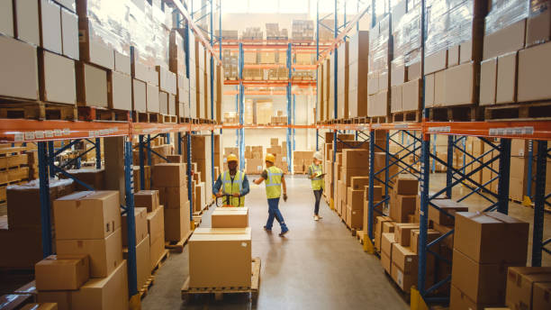 istock Retail Warehouse full of Shelves with Goods in Cardboard Boxes, Workers Scan and Sort Packages, Move Inventory with Pallet Trucks and Forklifts. Product Distribution Logistics Center. Elevated Shot 1284193247